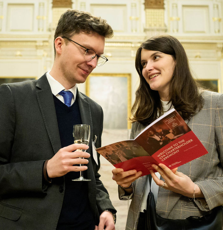Couple at charterholder ceremony looking at each other. The male is holding a glass of wine and the female is holding event program