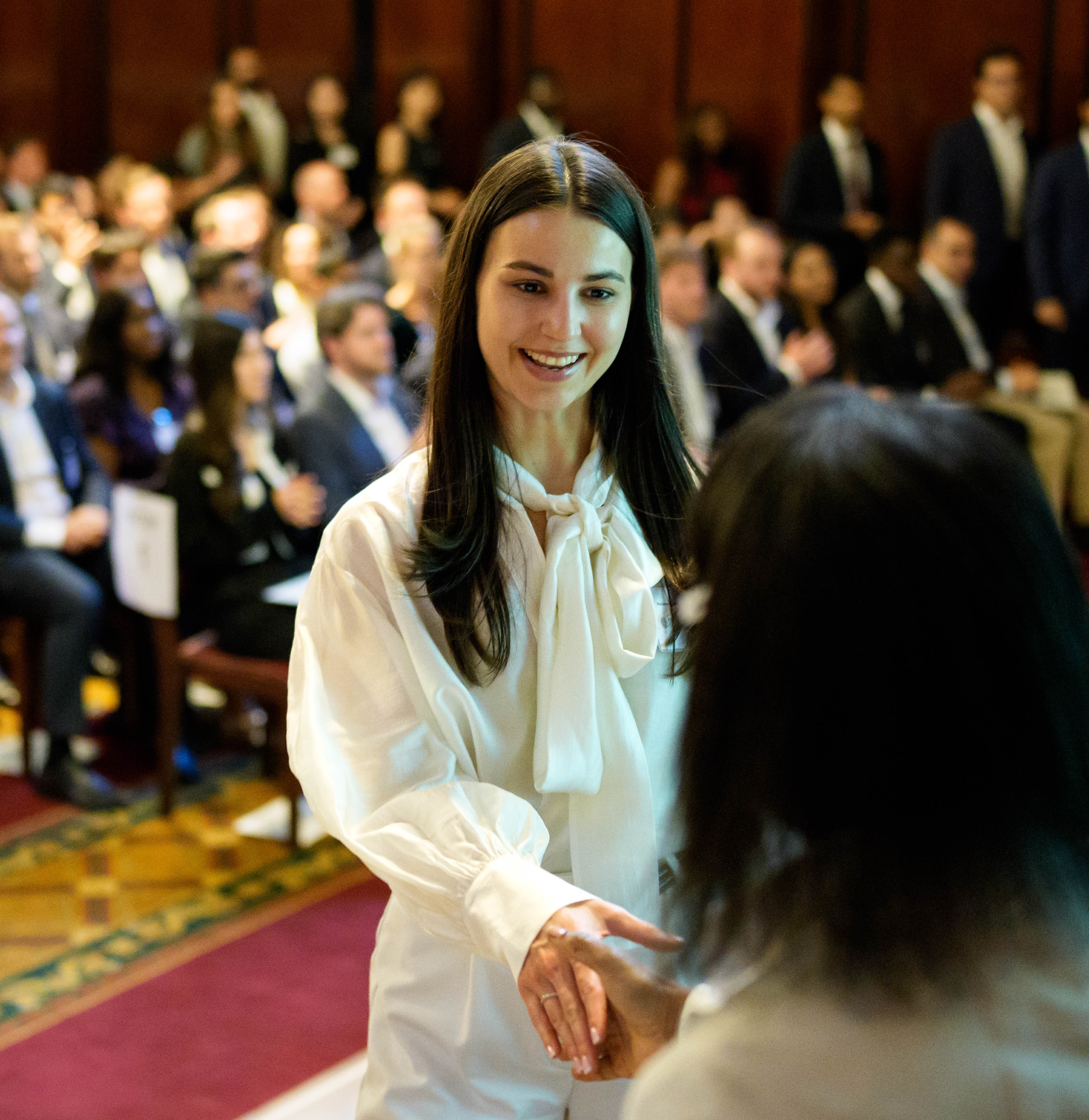 A CFA Charterholder graduate shakes hands with Sylvia Solomon, ASIP, CFA UK Board Chair, during a formal graudation ceremony, with an audience seated in the background.